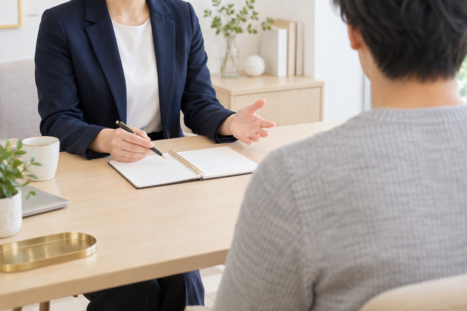 明るい相談室で、紺のジャケットのカウンセラーが机のノートを示しながら、グレーのニットの若い男性と話している様子
