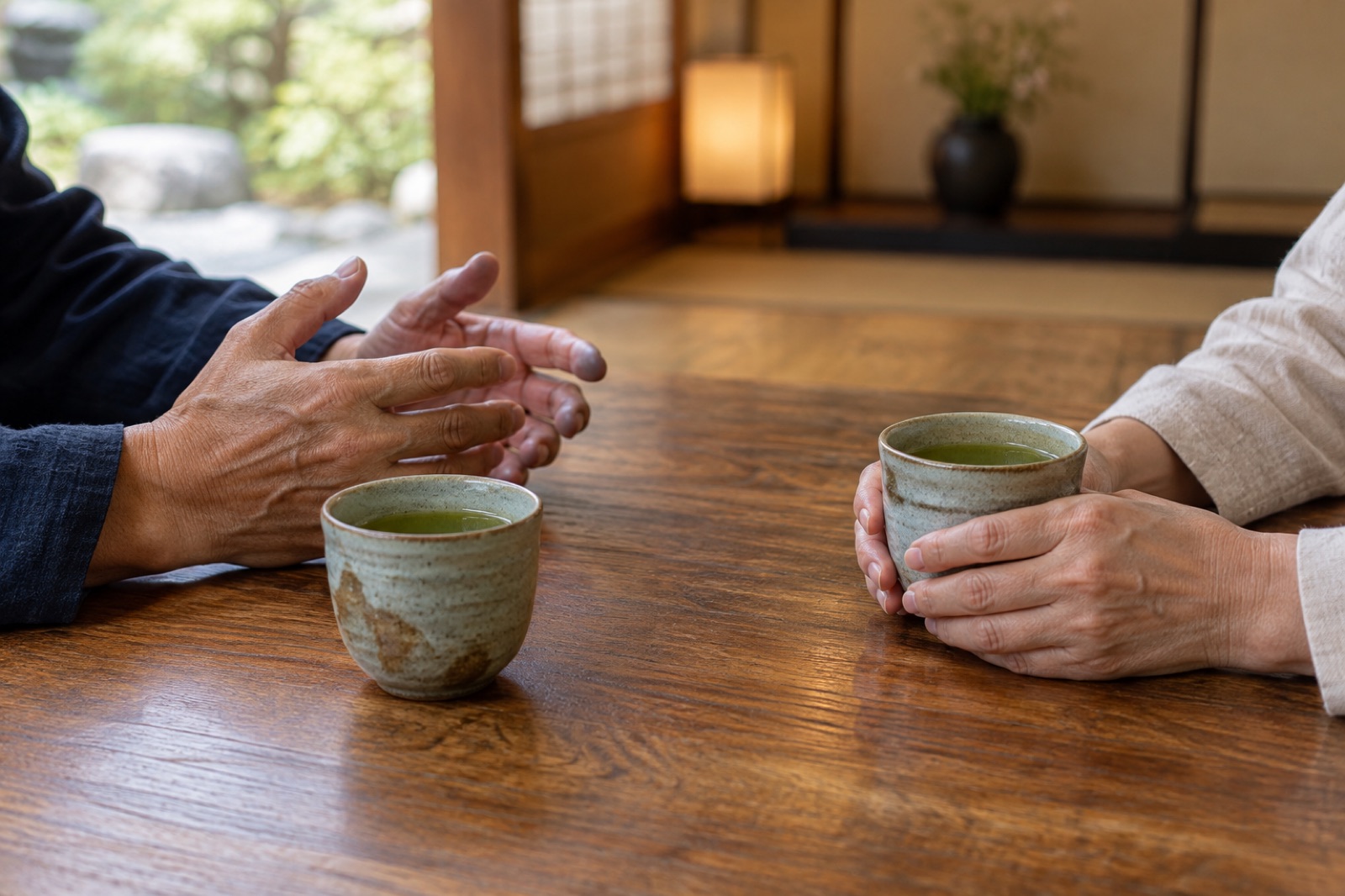 湯呑みを前に、身ぶりをまじえて話す人と、湯呑みを両手で包んで聞く人の手元