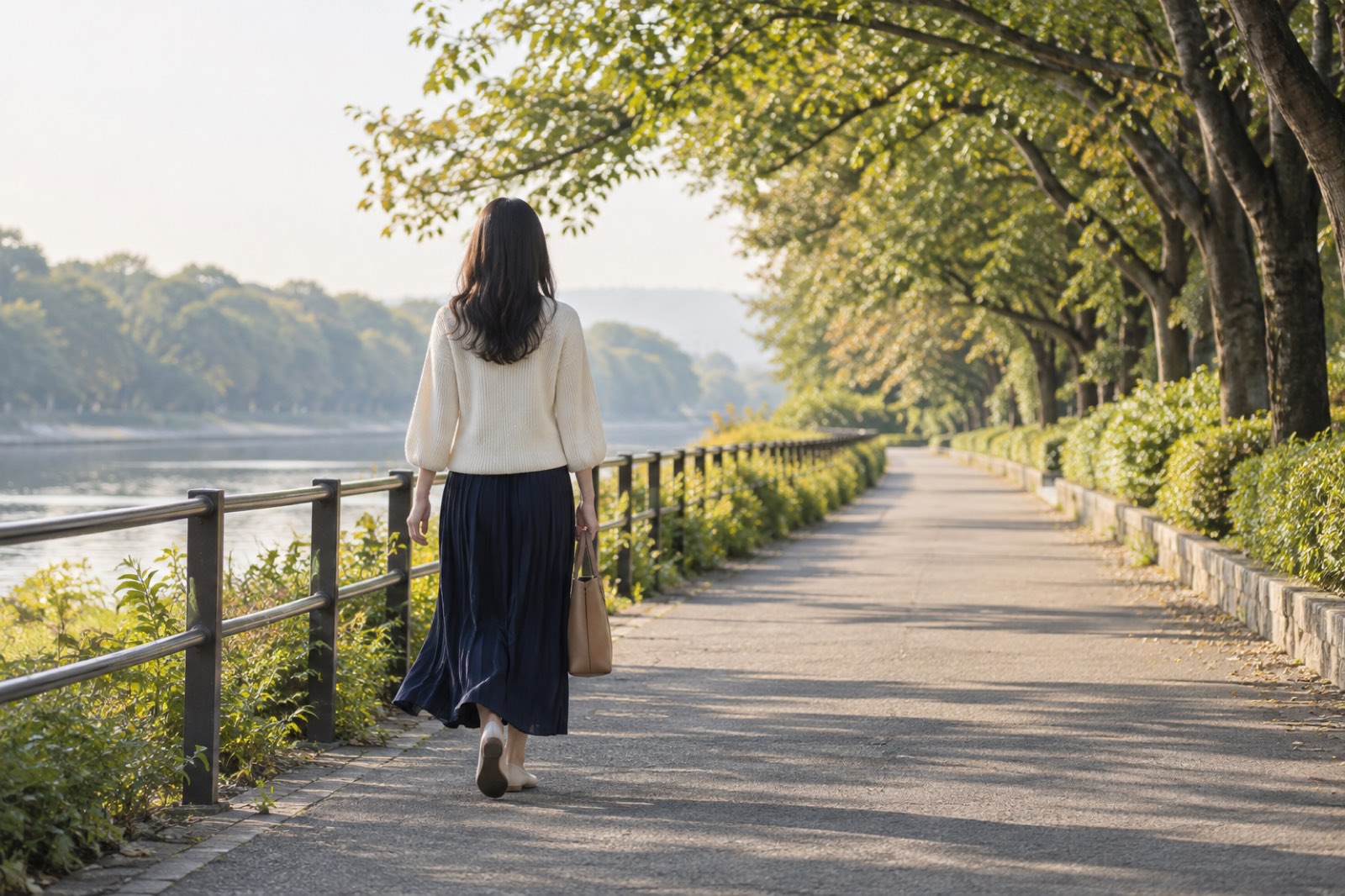 休日の朝、生成りのニットに紺のスカートで川沿いの並木道を歩く女性の後ろ姿