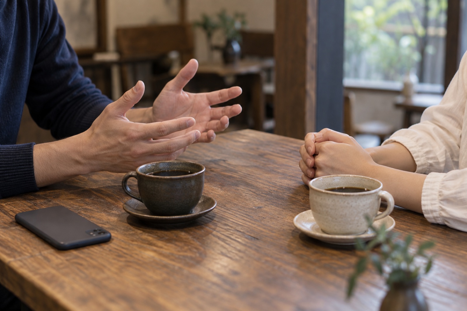 カフェのテーブルでスマートフォンを伏せ、向き合って話す二人の手元