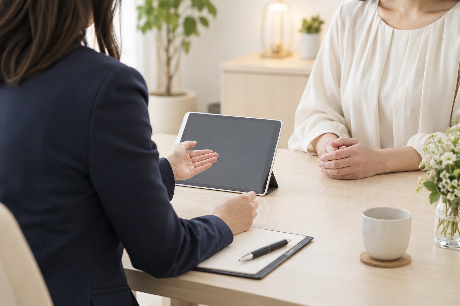 明るい相談室で、紺のジャケットのカウンセラーが机のタブレットを示しながら、手を重ねて座る女性と話している様子