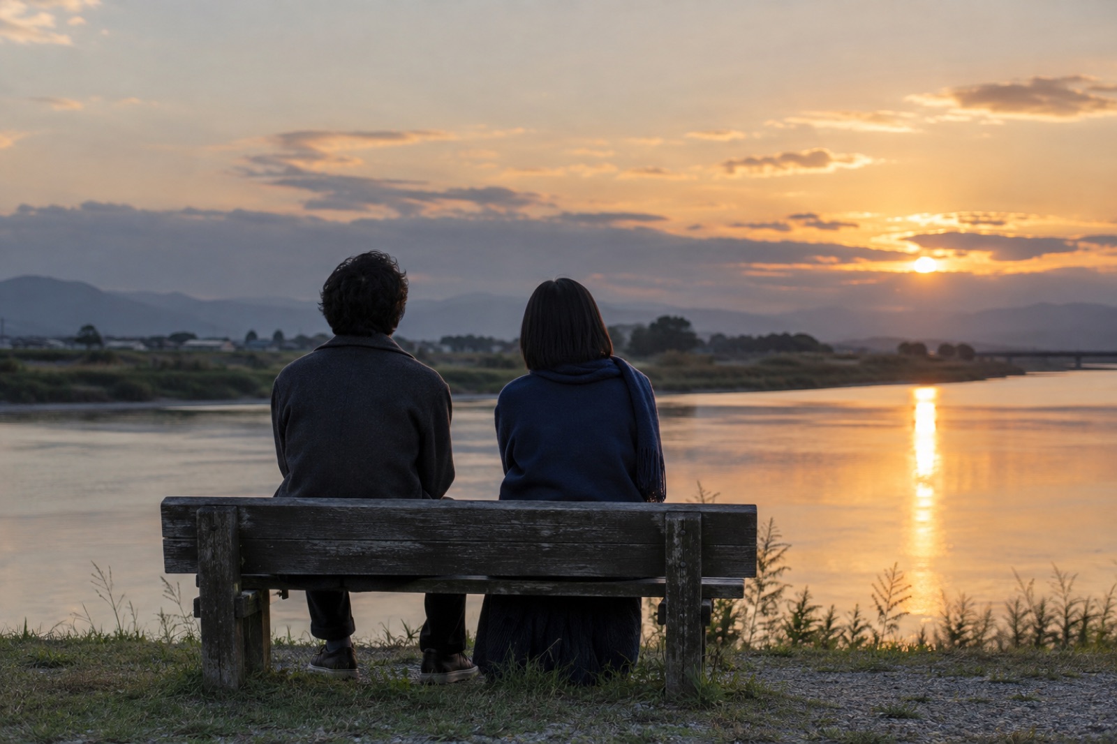 夕暮れの川辺のベンチに、くつろいで並んで座る二人の後ろ姿