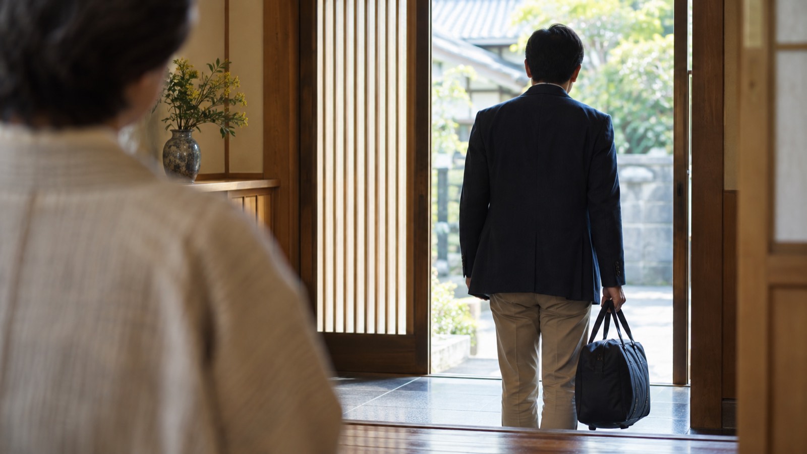 実家の玄関から帰っていく大人の子の後ろ姿と、見送る親の肩