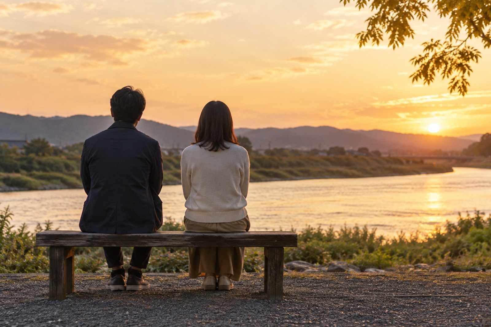 夕暮れの川辺のベンチに並んで座る二人の後ろ姿